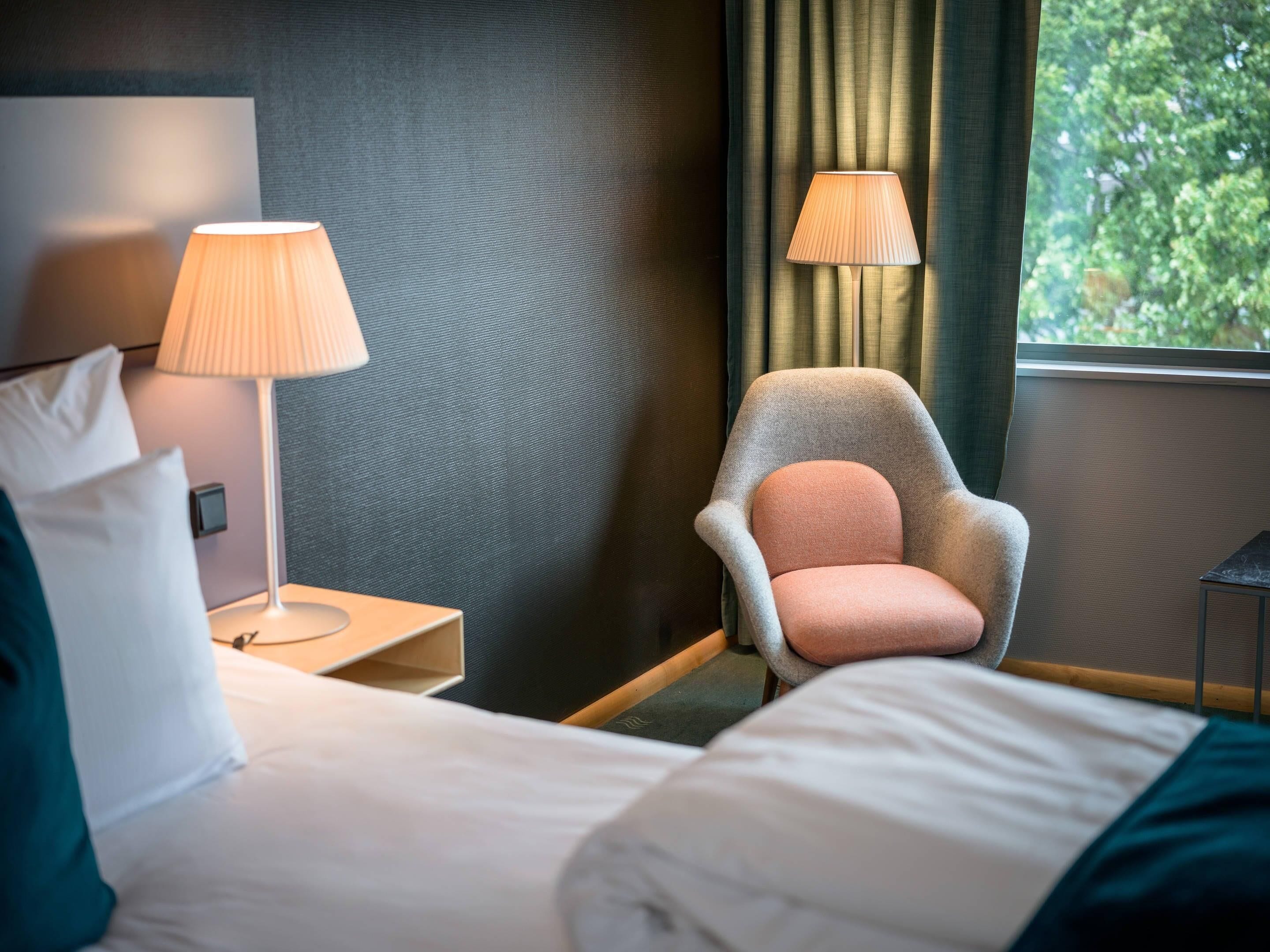 Una habitación de hotel con una cama, una silla, dos lámparas y un ventanal con cortinas.