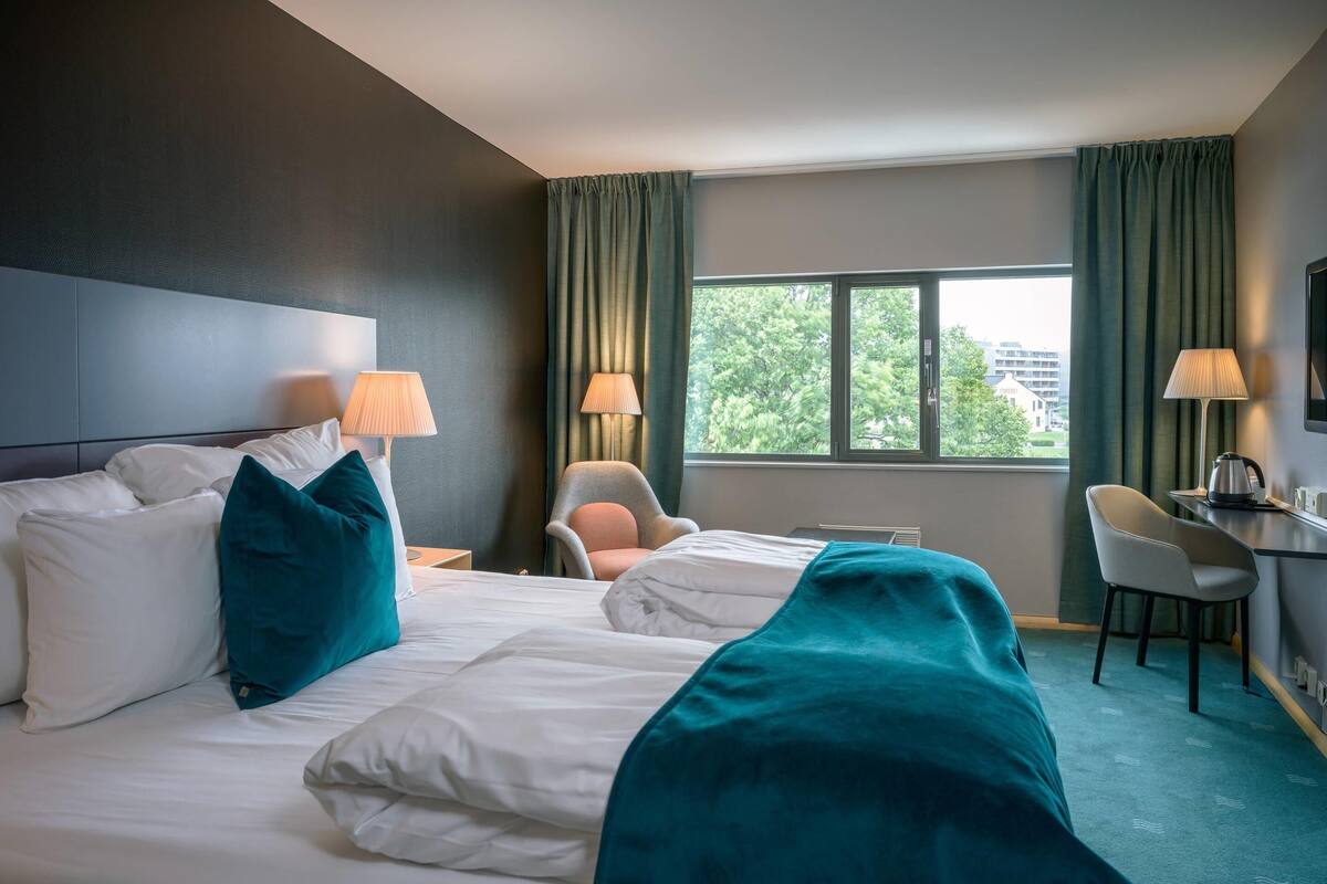 A hotel room with a bed, two chairs, a desk, and a view of trees through the window.