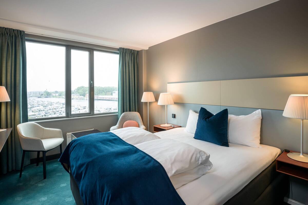 A hotel room with a large bed, two chairs, a view of the water, and a cityscape.
