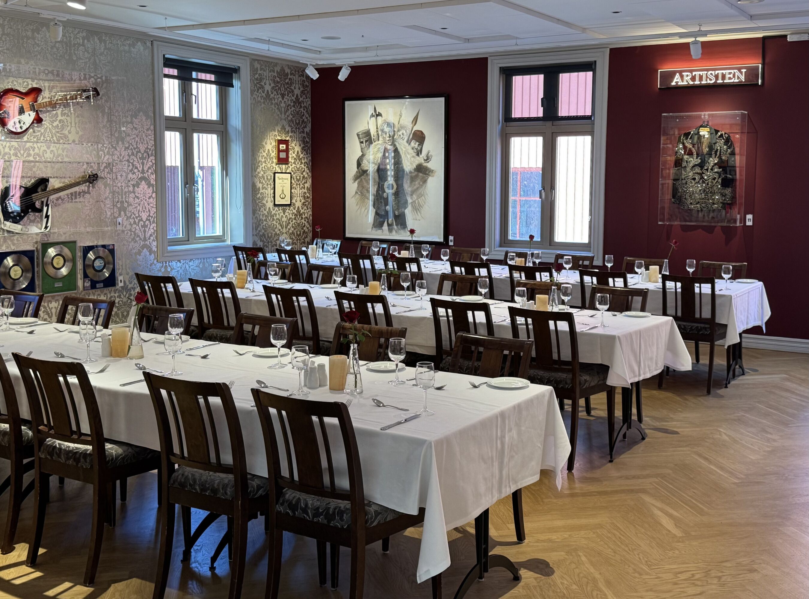 A dining area with tables set for service, framed artwork on the walls, and a sign reading 'ARTISTEN'.