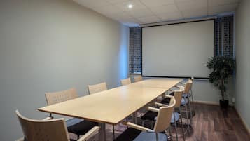 A conference room with a long table, chairs, a projector screen, and a potted plant.