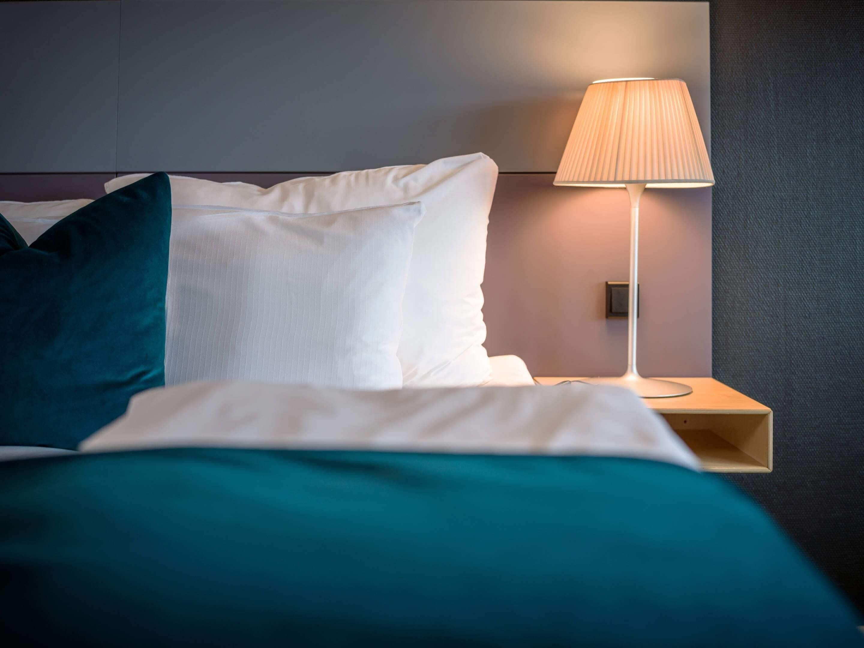 A neatly made bed with white and teal bedding, a bedside lamp, and a wall-mounted control panel.