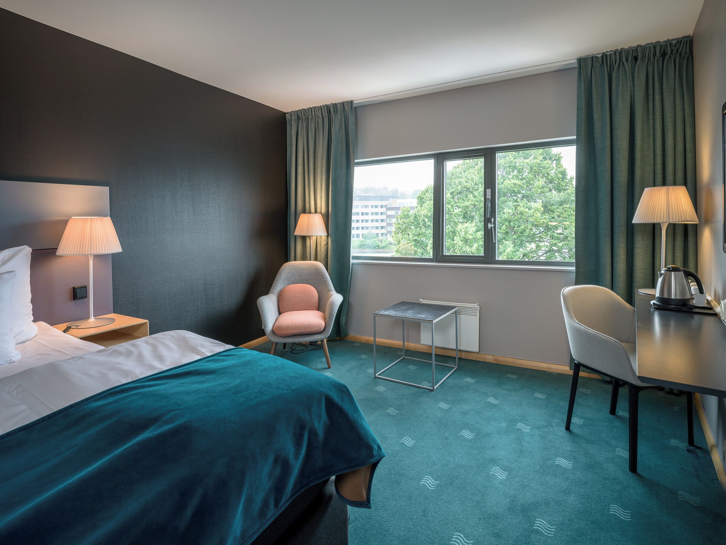 A hotel room with a bed, two chairs, a small table, a desk, and a window with curtains.