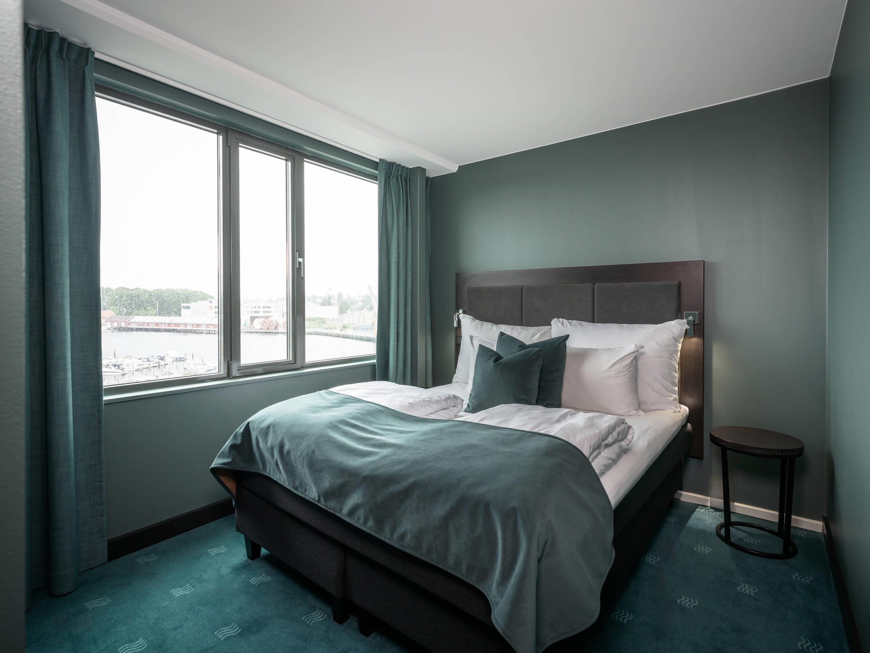 Habitación de hotel con una cama grande, una mesita auxiliar pequeña y vistas de la ciudad a través de un ventanal amplio.