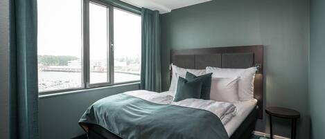 Habitación de hotel con una cama grande, una mesita auxiliar pequeña y vistas de la ciudad a través de un ventanal amplio.