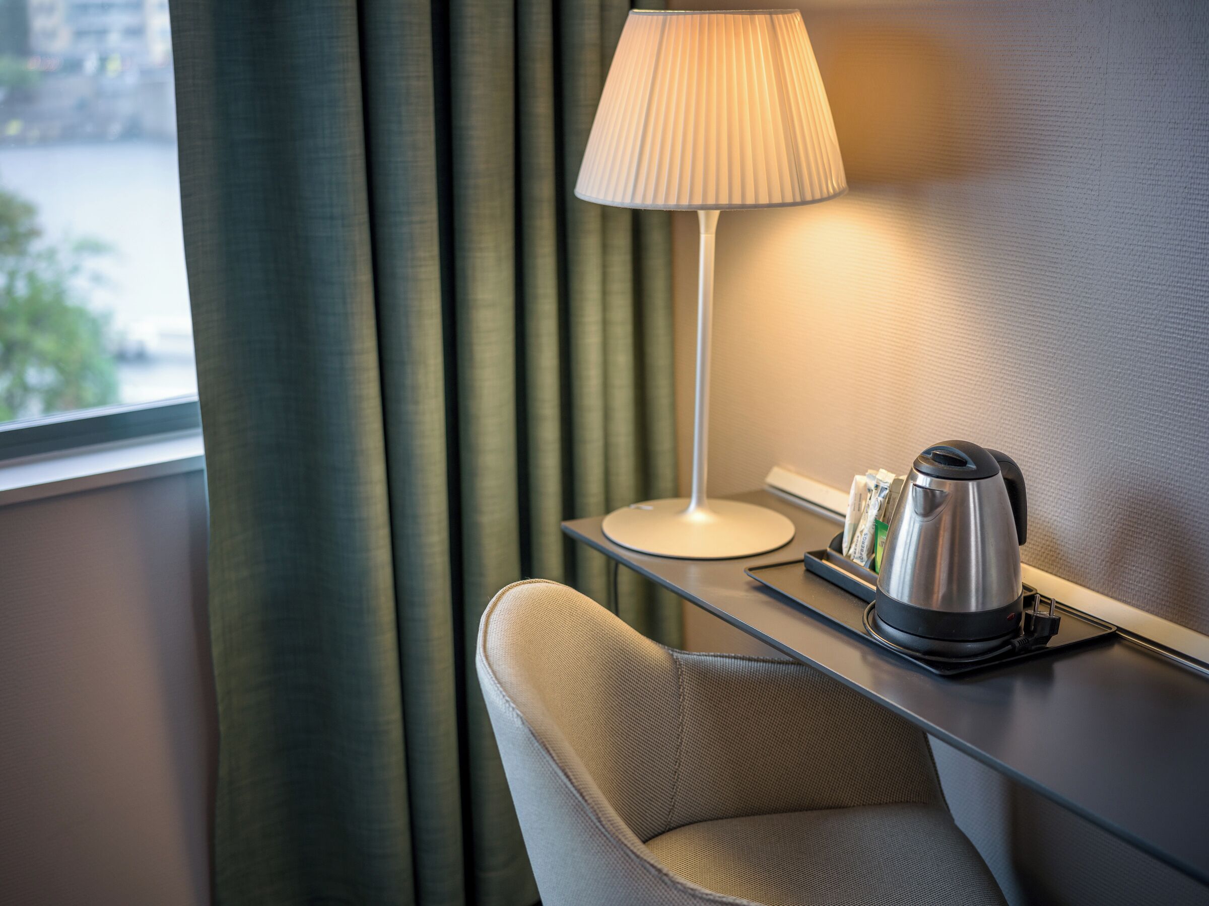 Habitación de hotel con un escritorio moderno, una silla, una lámpara y una tetera.