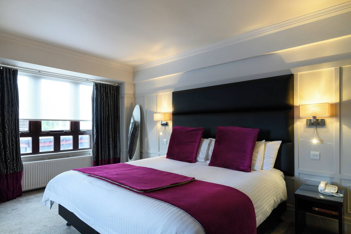 A hotel room with a large bed, two bedside lamps, a nightstand with a phone, and a window with curtains.