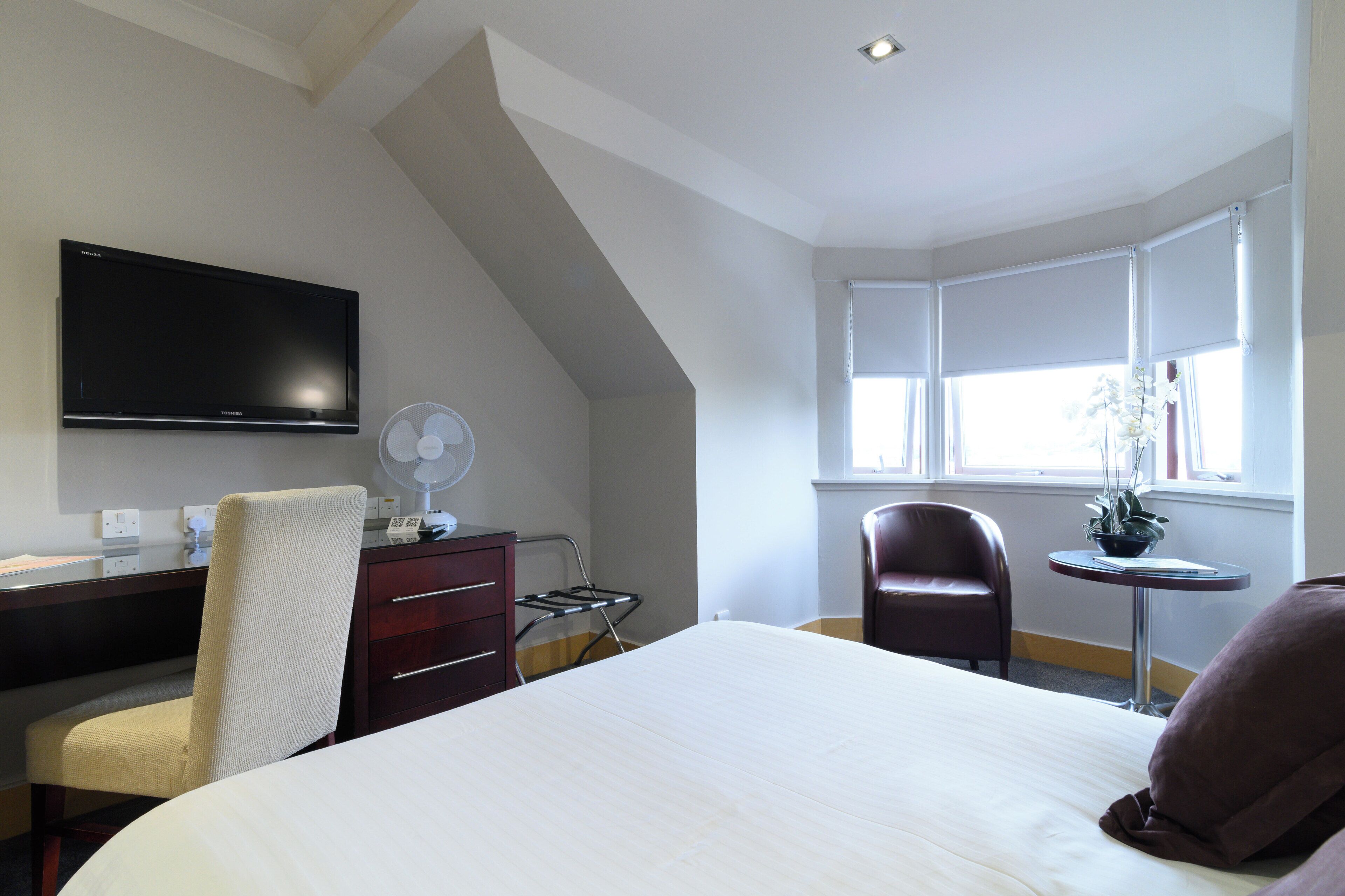 A hotel room with a bed, a desk with a flat-screen TV, a chair, a small table with a plant, and a window with curtains.