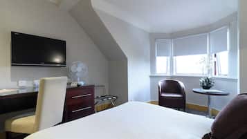 A hotel room with a bed, a desk with a flat-screen TV, a chair, a small table with a plant, and a window with curtains.