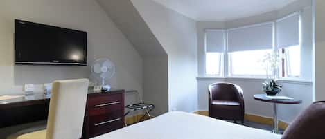 A hotel room with a bed, a desk with a flat-screen TV, a chair, a small table with a plant, and a window with curtains.