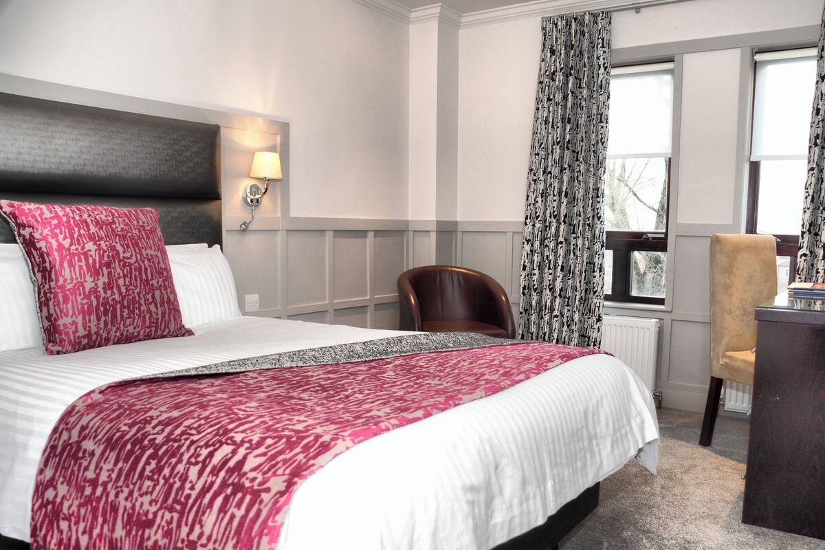 A hotel room with a large bed, a desk, a chair, a lamp, and a window with curtains.