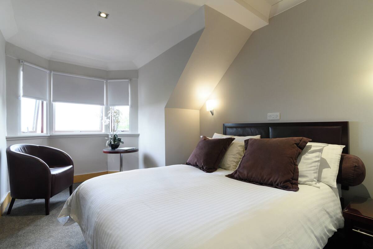 A bedroom with a large bed, a chair, a small table, and a window with blinds.