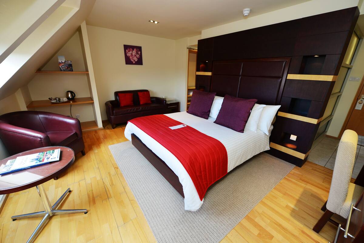 A bedroom with a large bed, a red and white bedspread, a dark headboard, a red leather chair, a small table with a magazine, and a wooden floor.