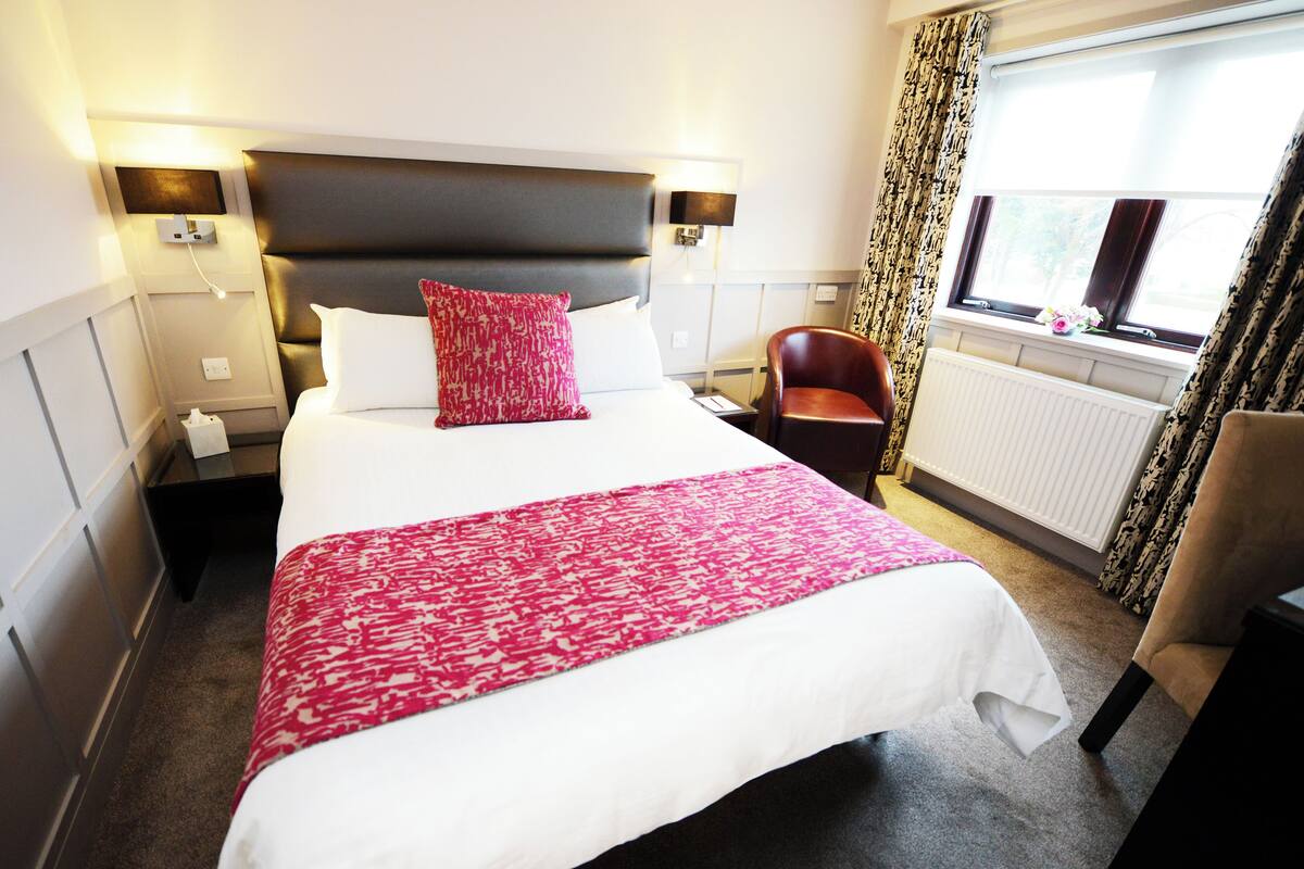 A hotel room with a large bed, a red and white patterned throw, a dark headboard, a nightstand with a lamp, a chair, and a window with curtains.