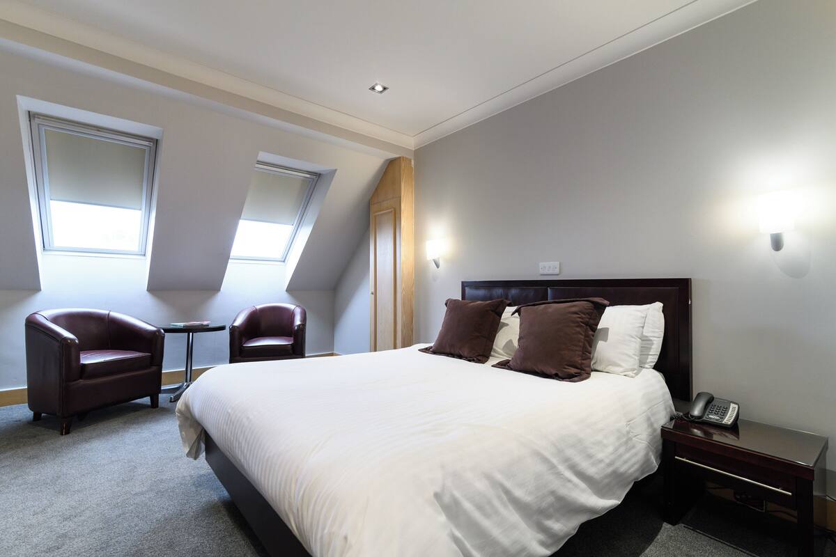 A hotel room with a large bed, two armchairs, a small table, and a nightstand with a phone.