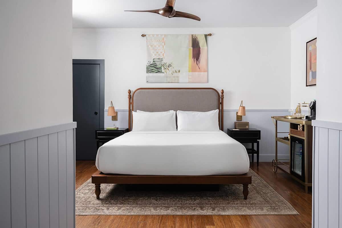 A bedroom with a wooden bed, a ceiling fan, a nightstand with a lamp, a small table with a lamp, and a wall art piece.