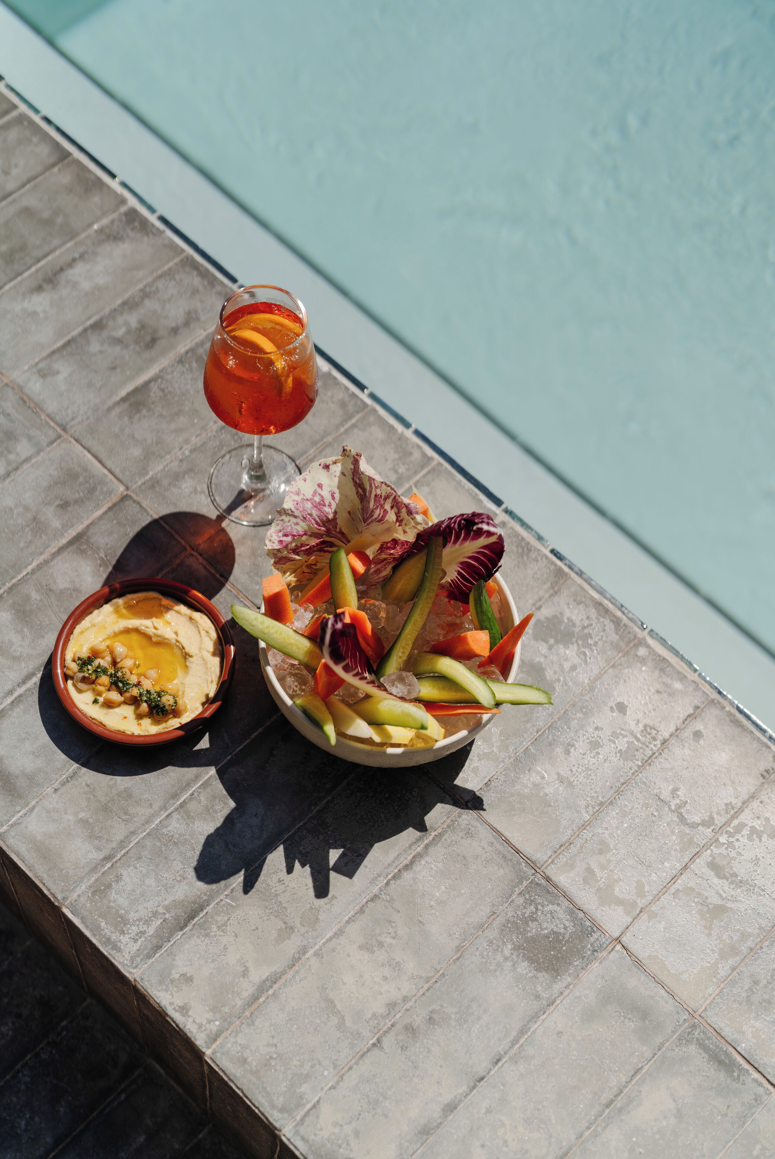 Ein Tisch mit Hummus, einem Glas rotem Getränk und einem Obstsalat am Pool.