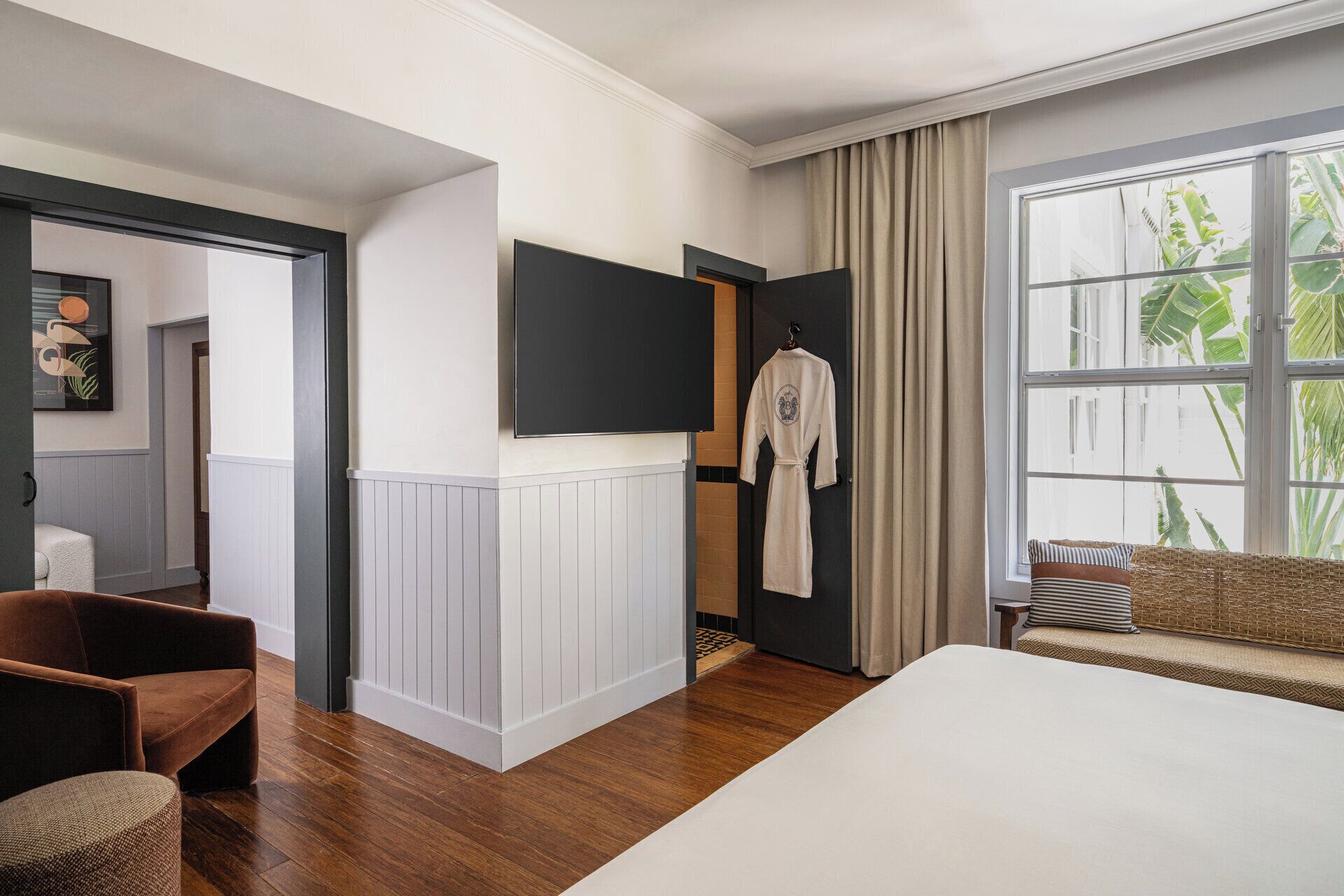 A hotel room with a large bed, a flat-screen TV mounted on the wall, a robe hanging on a hook, and a window with a view of greenery.