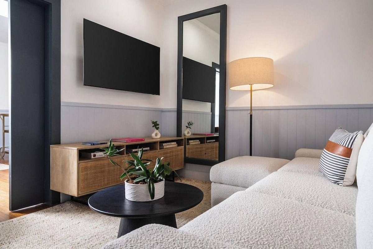 A modern living room with a sofa, a coffee table with a plant, a side table, a lamp, and a large mirror.