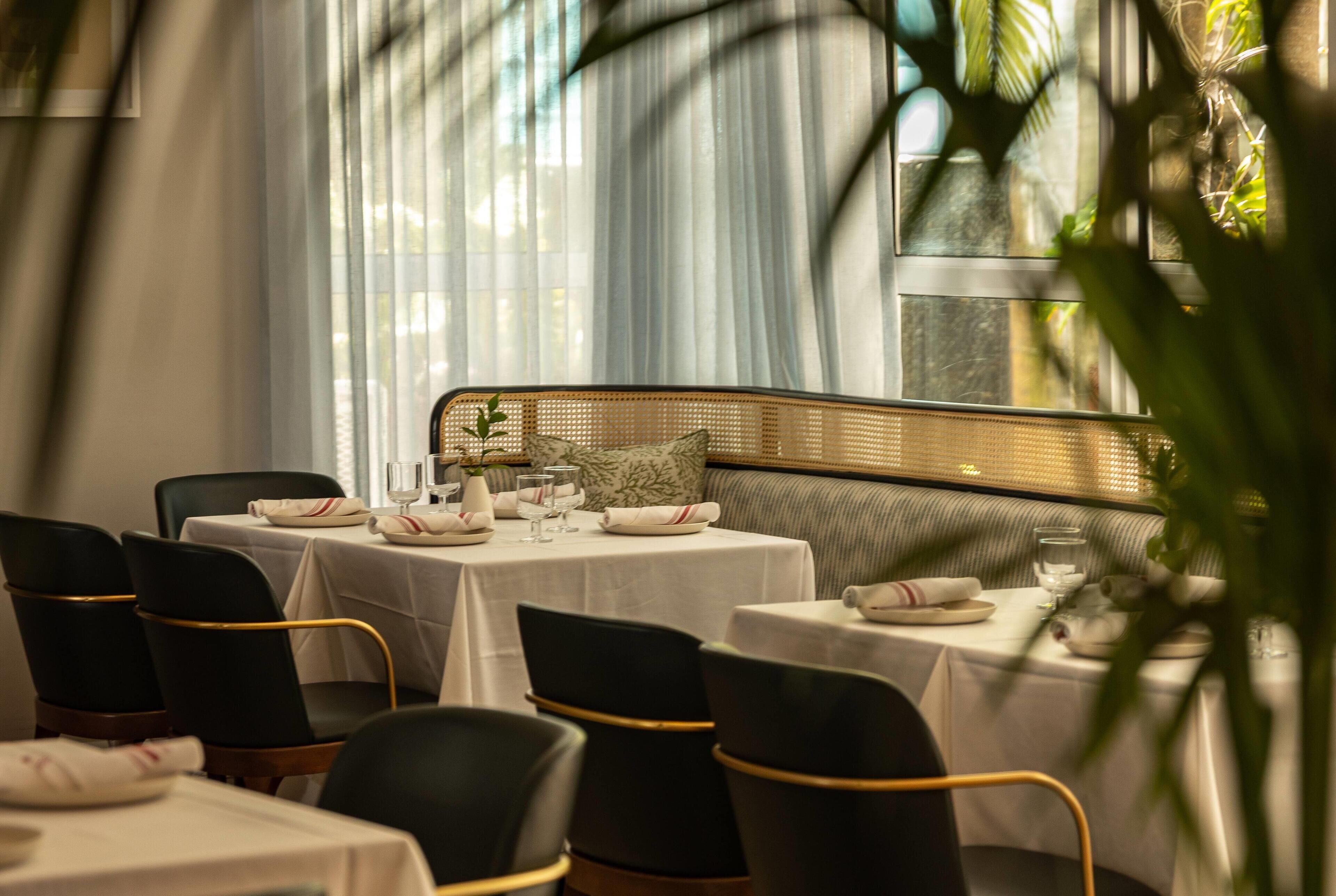 A dining area with tables set for four, each with a white tablecloth, black chairs with gold accents, and a central woven bench.