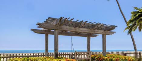 A wooden pergola with a swing, surrounded by colorful bushes and a wooden fence, overlooking a beach.