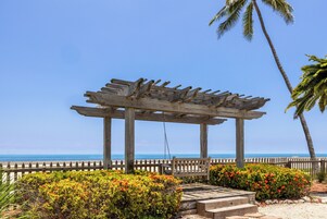 Una pérgola de madera con un columpio, rodeada de arbustos coloridos y una cerca de madera, con vista a una playa.