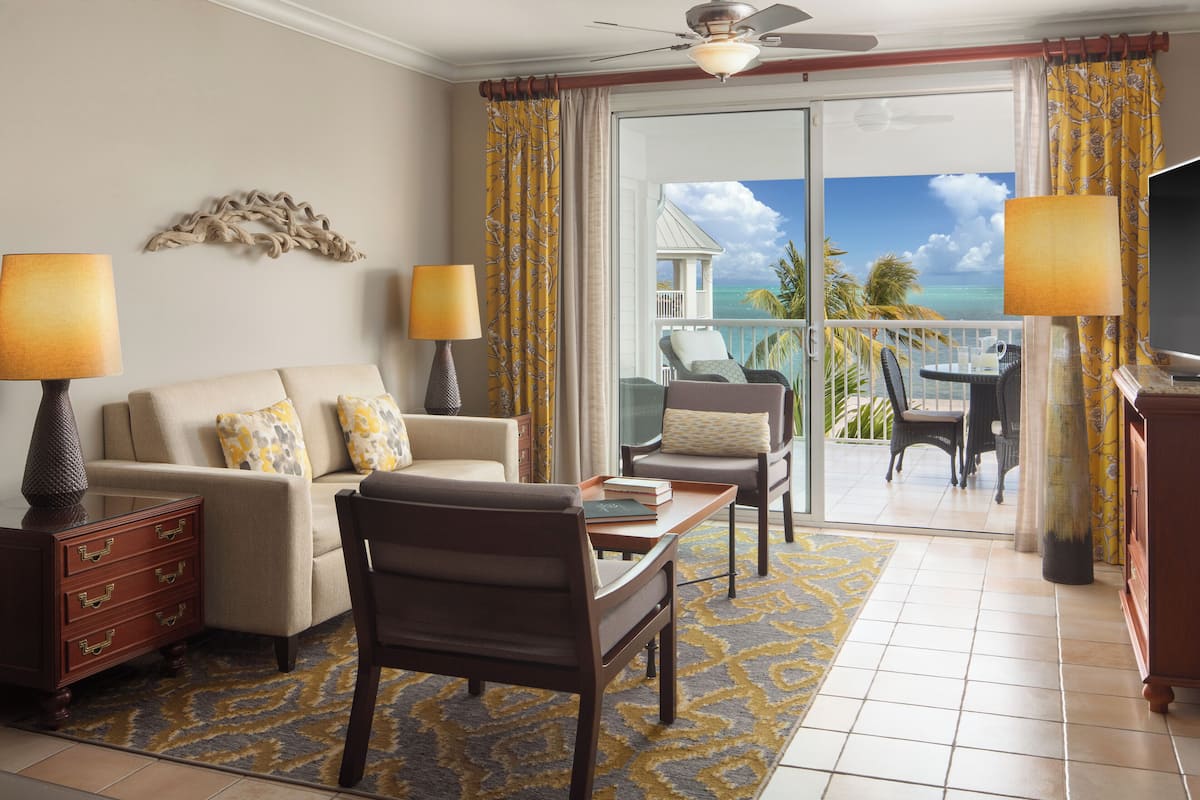 A living room with a sofa, armchairs, a coffee table, and a TV. There are two lamps, a decorative wall piece, and a view of the ocean through a sliding door.
