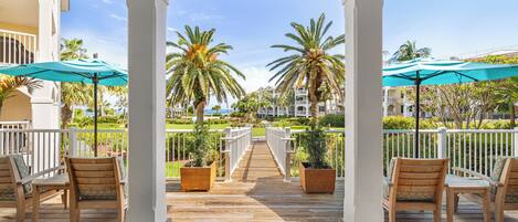 A wooden deck with chairs and umbrellas, leading to a white fence and palm trees.
