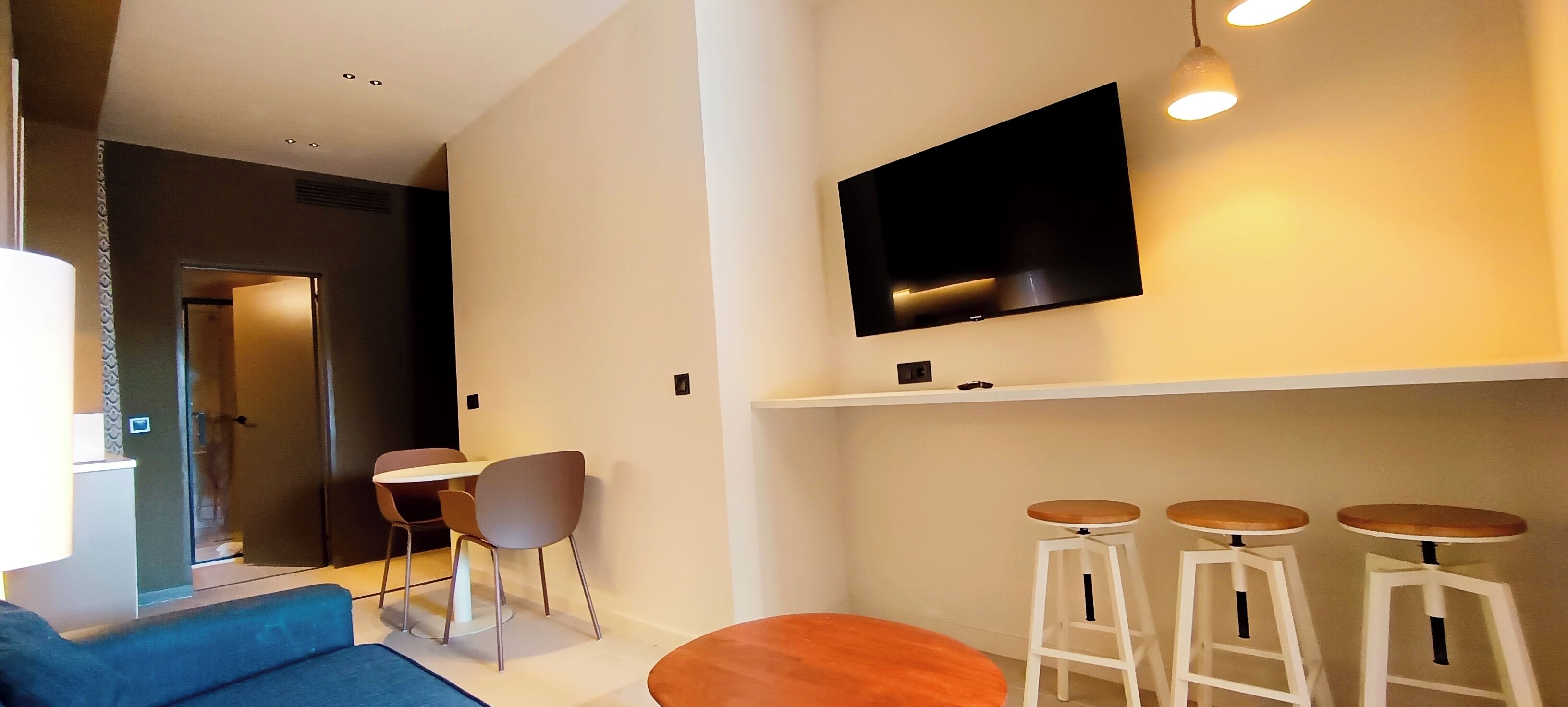 Een moderne woonkamer met een flatscreen televisie aan de muur, een blauwe bank, een kleine ronde tafel en drie witte barstoelen.