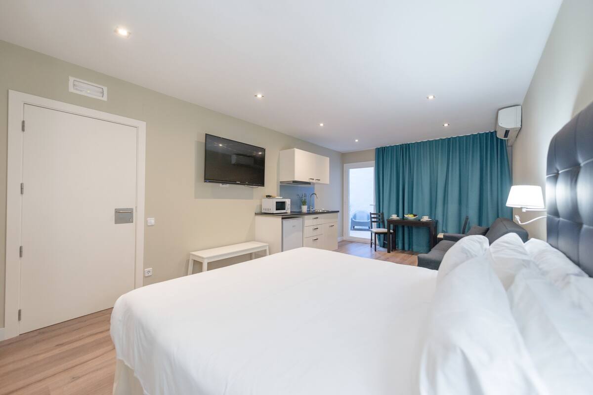 Une chambre d’hôtel avec un grand lit, un téléviseur fixé au mur, une kitchenette et une vue sur l’extérieur par une fenêtre munie de rideaux.