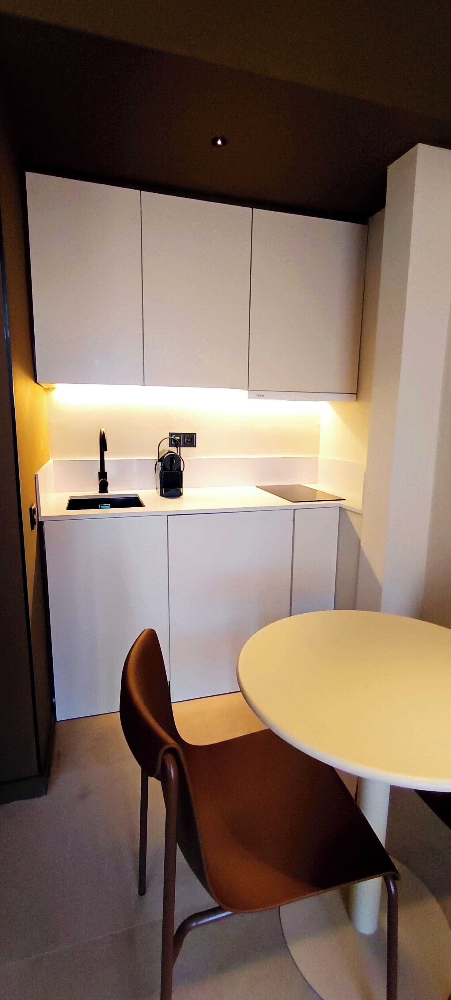 Cocina compacta con armarios blancos, encimera blanca, un lavamanos y una mesa redonda con una silla marrón.