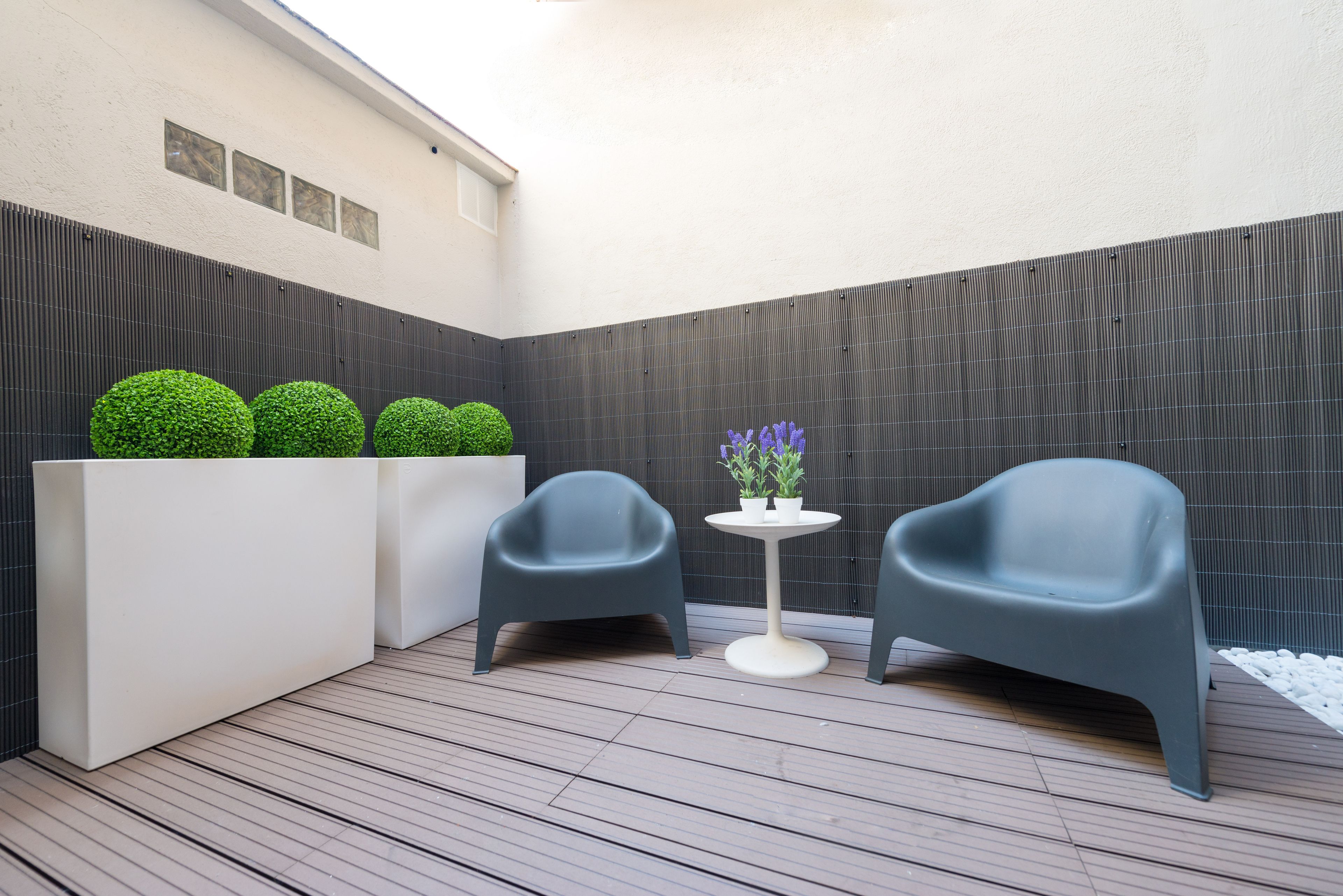 Un espace extérieur moderne aménagé avec deux chaises et une petite table, entouré d’une terrasse en bois et d’un bac à fleurs blanc contenant des arbustes verts.