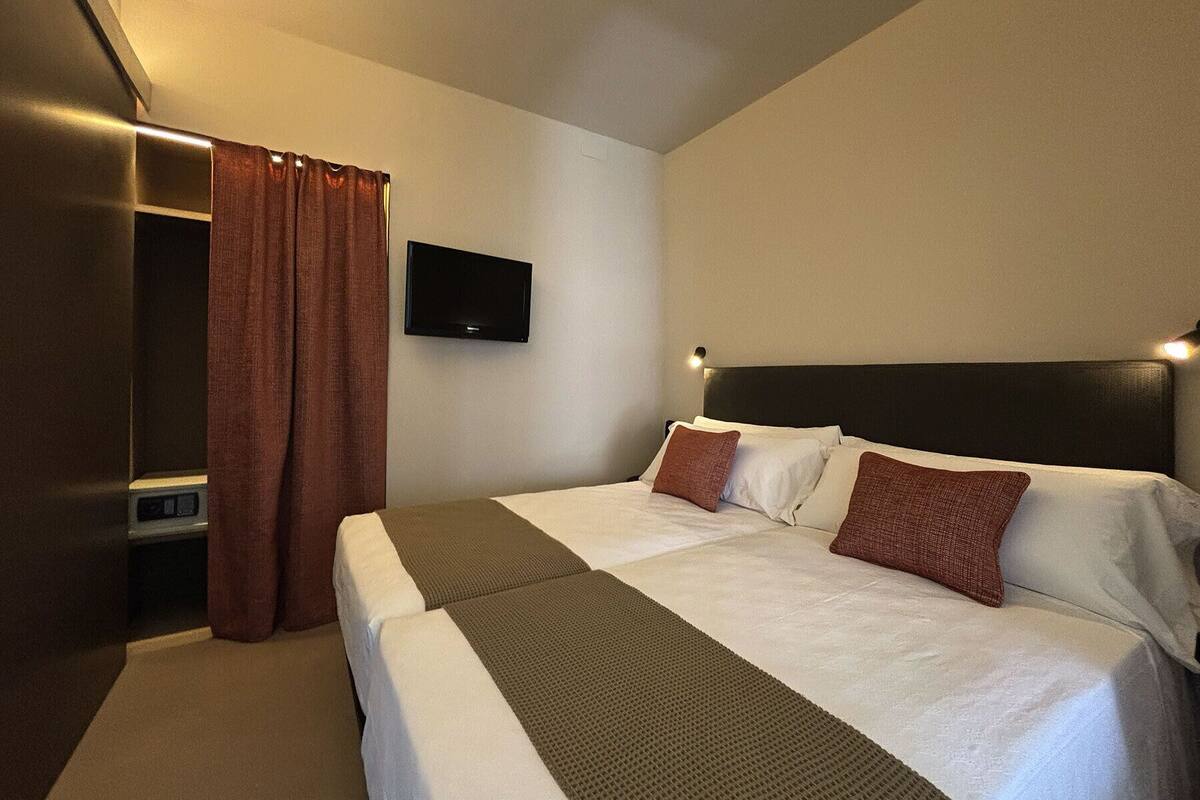 Une chambre d’hôtel avec deux lits, un téléviseur fixé au mur et un rideau sur le côté gauche.
