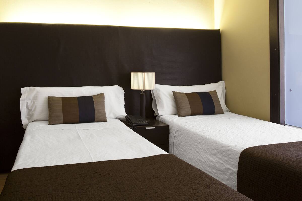 Une chambre d’hôtel avec deux lits simples, une table de chevet avec une lampe et une vue sur l’extérieur.
