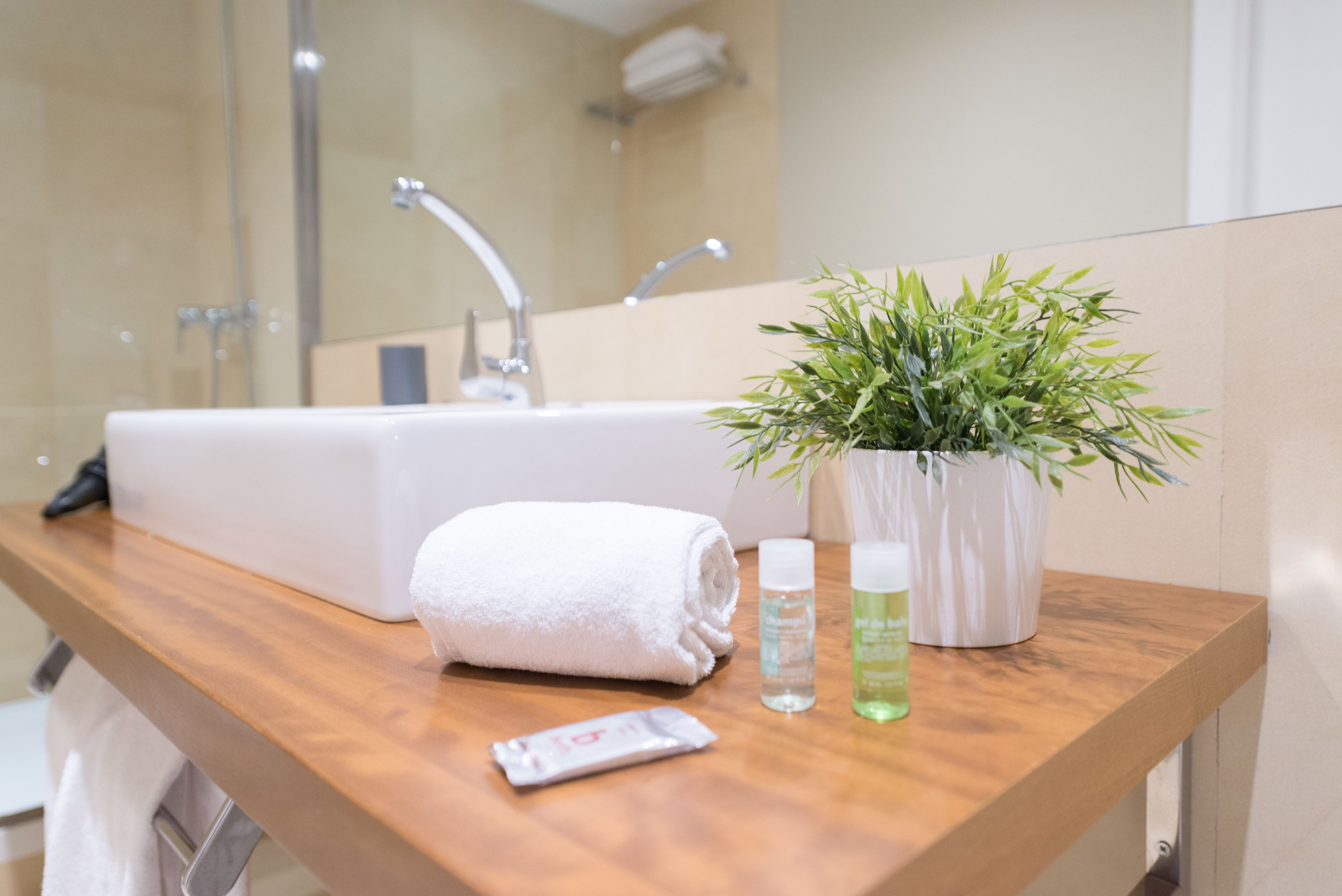Un baño con tocador de madera, un lavamanos blanco, una toalla, una planta en maceta y dos botellas de loción.
