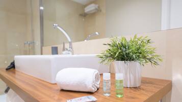 Un baño con tocador de madera, un lavamanos blanco, una toalla, una planta en maceta y dos botellas de loción.