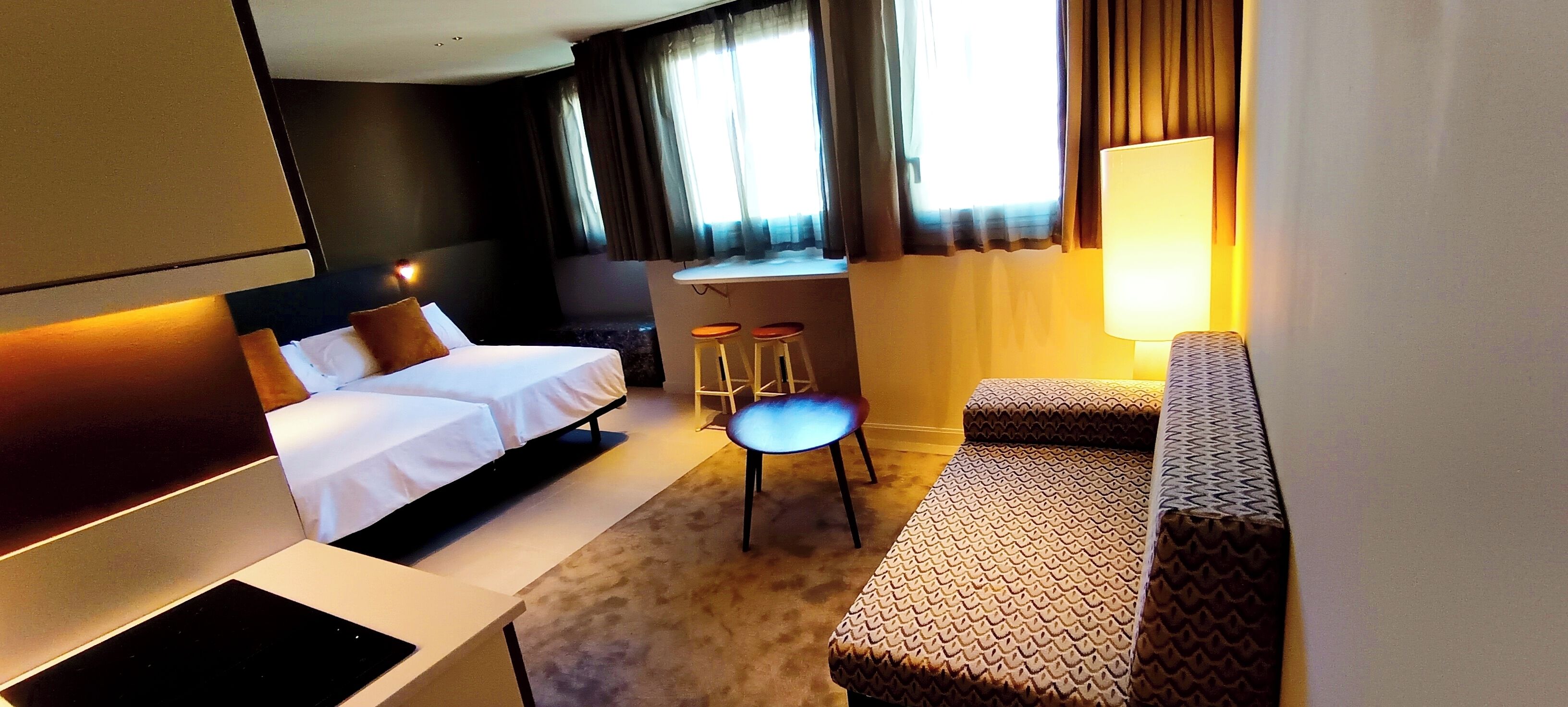 A hotel room with a bed, a desk with two stools, a sofa, and a lamp.