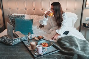 Quarto de hotel com cama, um laptop sobre o travesseiro, um tabuleiro com itens de pequeno-almoço e um copo de sumo de laranja.