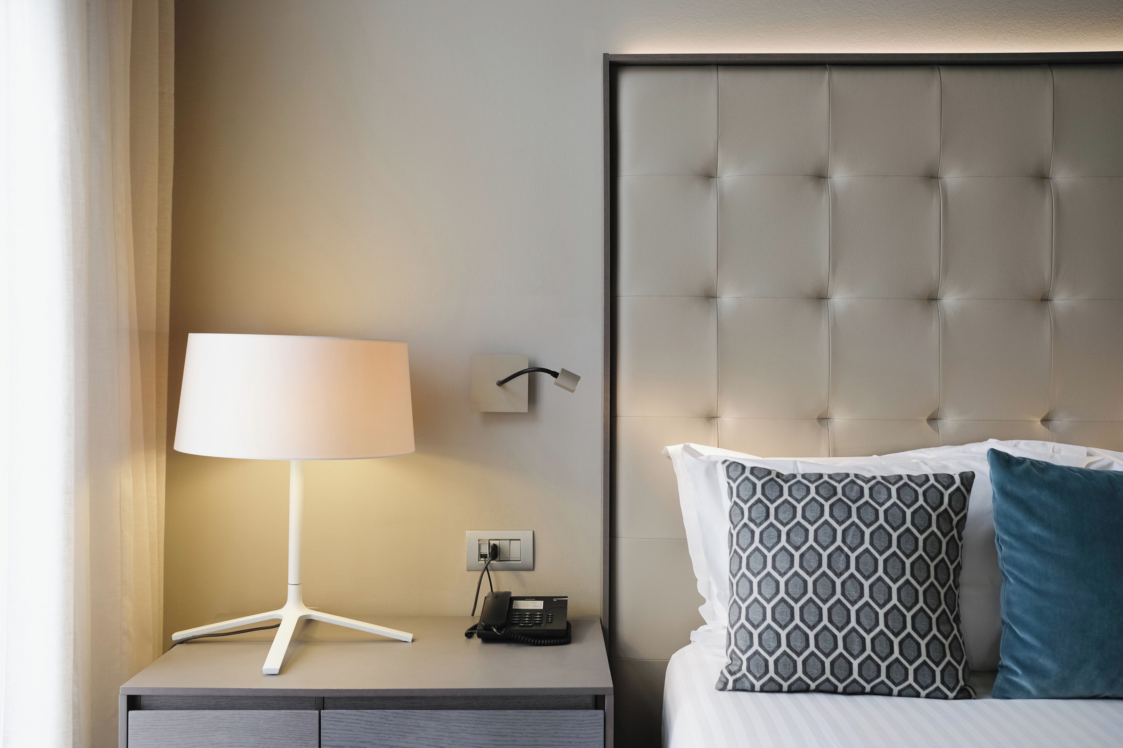 Una cama bien hecha con almohadones estampados, una lámpara de noche y un teléfono sobre una mesita.