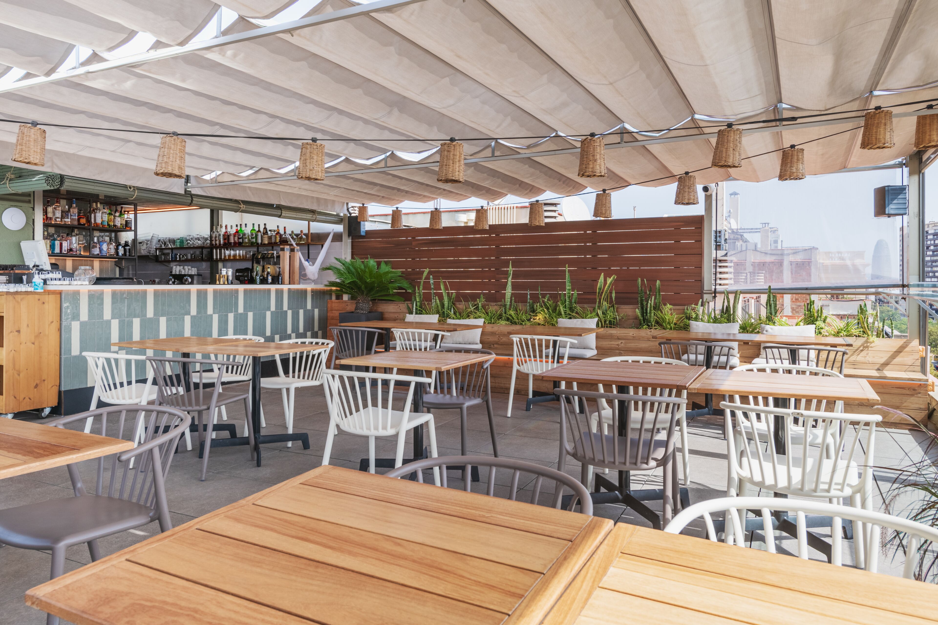 An outdoor café with wooden tables and white chairs, a bar area with shelves of bottles, and a view of the city through large windows.