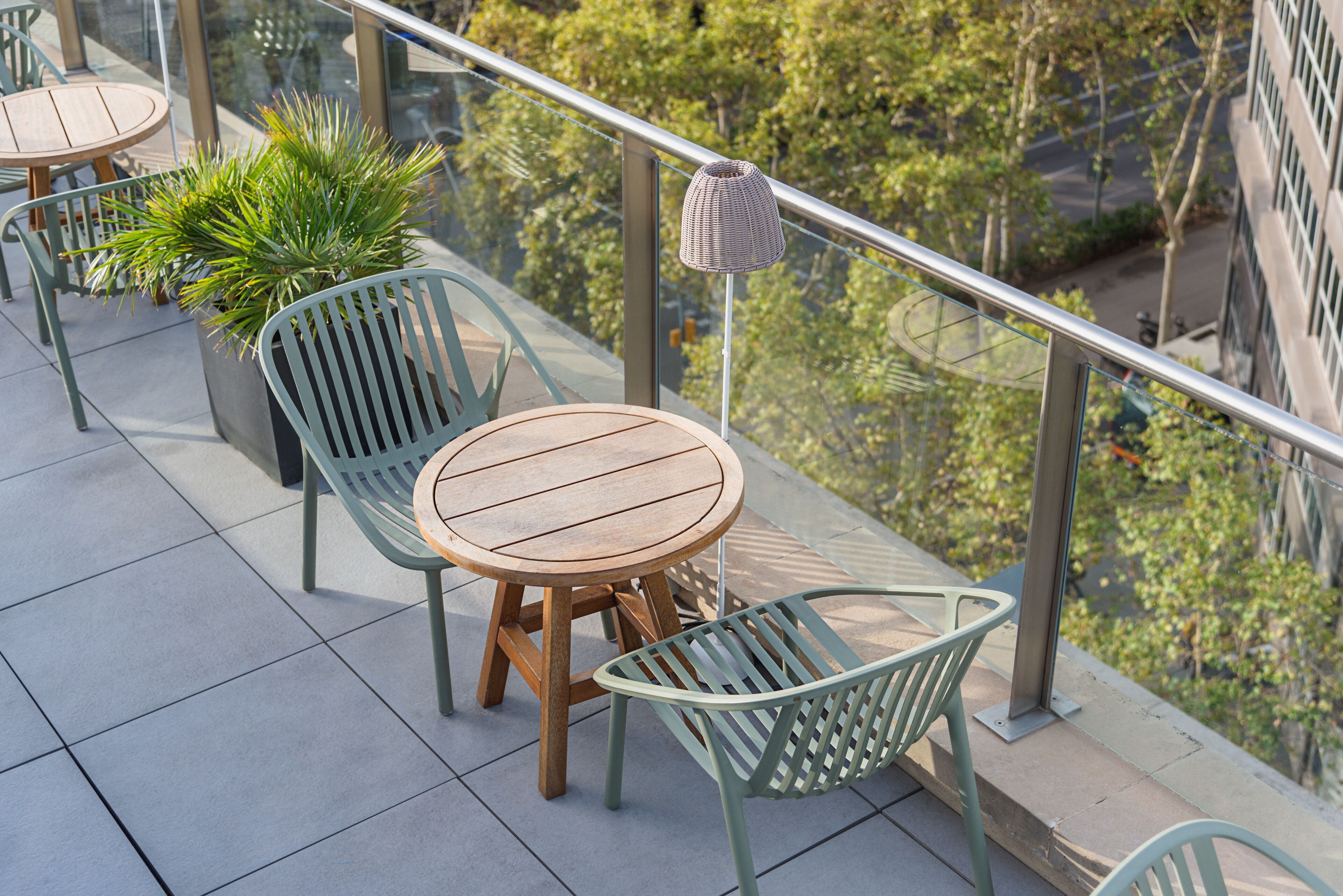 A balcony with a round wooden table and two green chairs, a potted plant, and a hanging lamp.