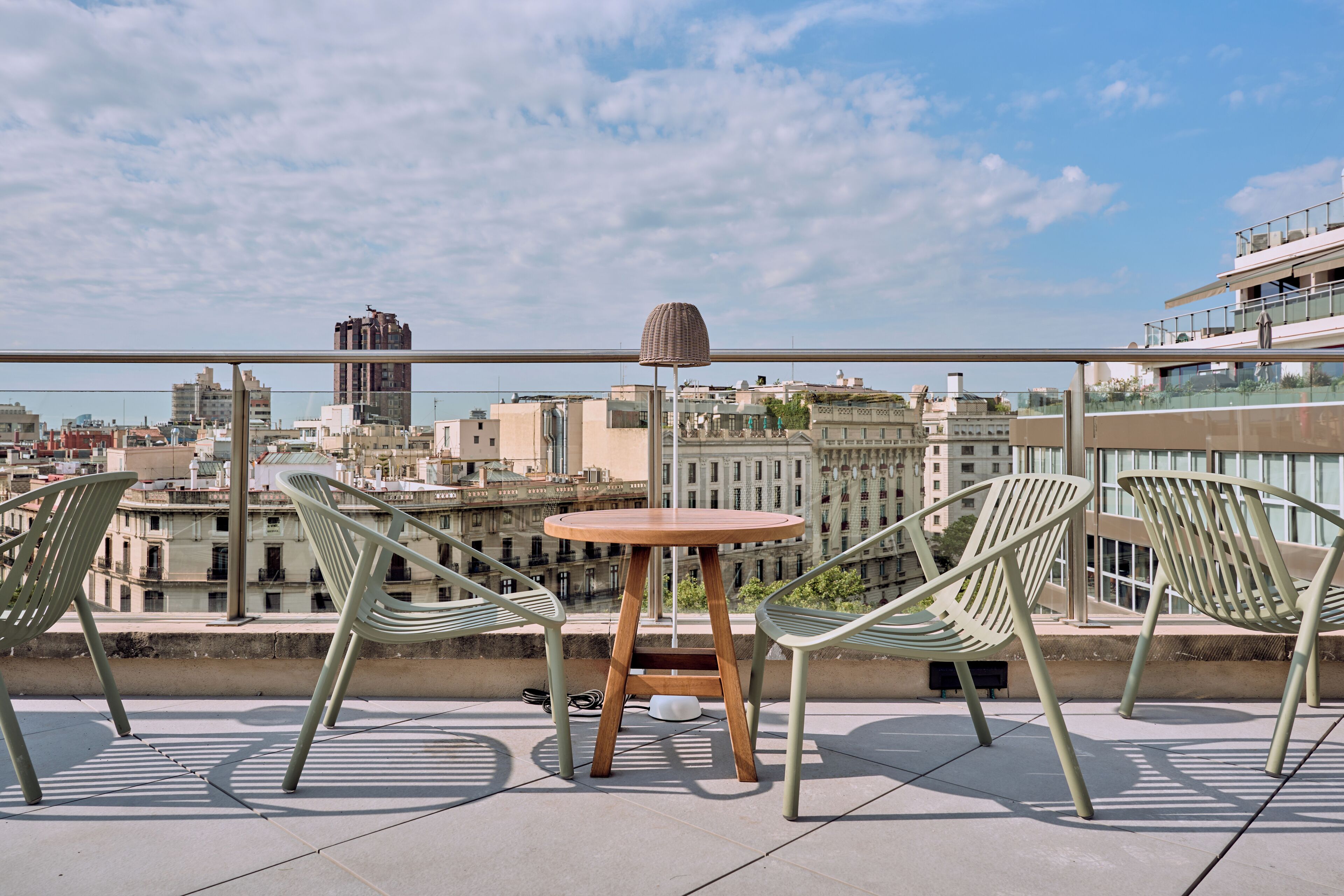 A rooftop terrace with a round table and two chairs, overlooking a cityscape with buildings and a clear sky.