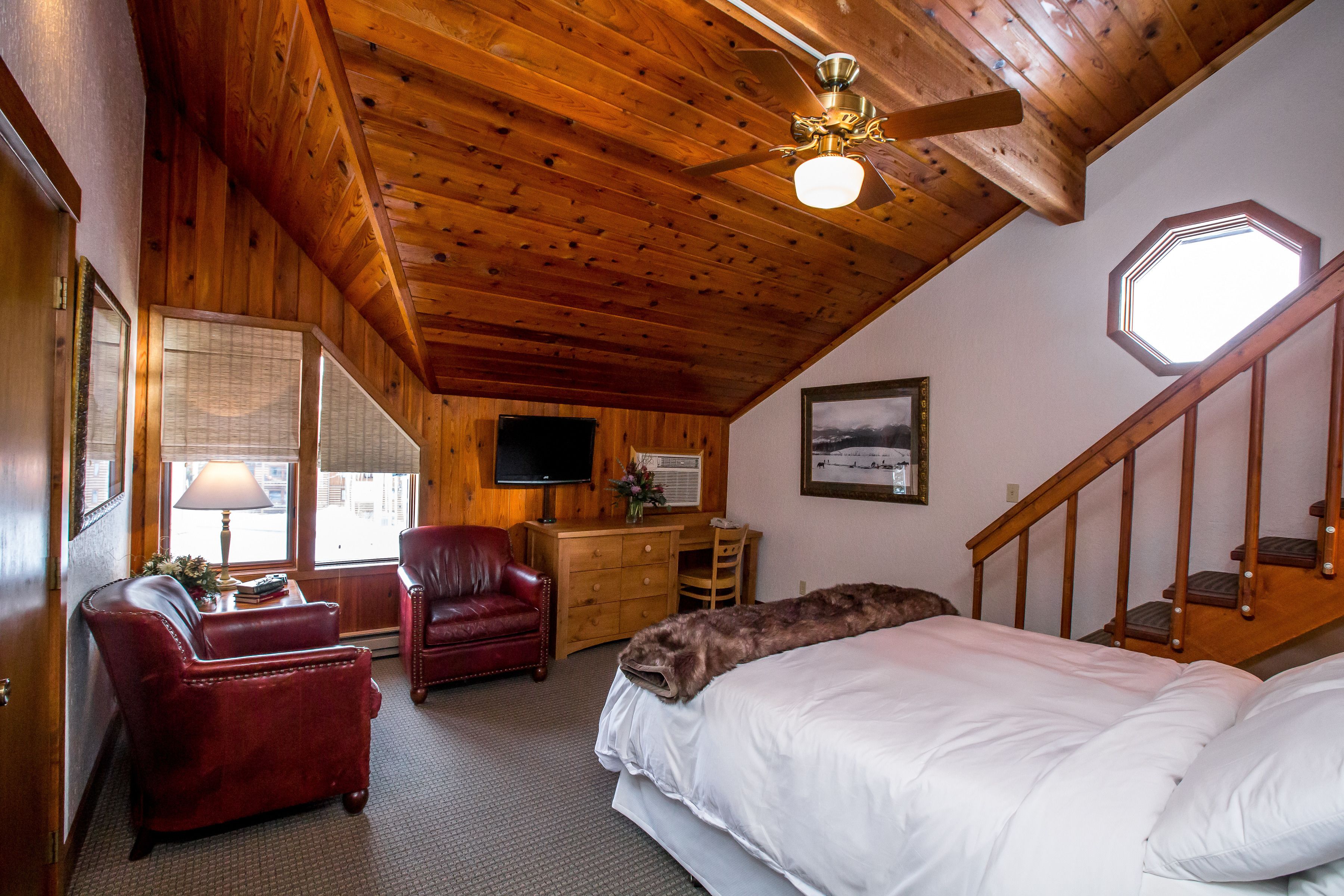 A room with a wooden ceiling, a bed, a desk with a chair, a red leather chair, and a television on the wall.