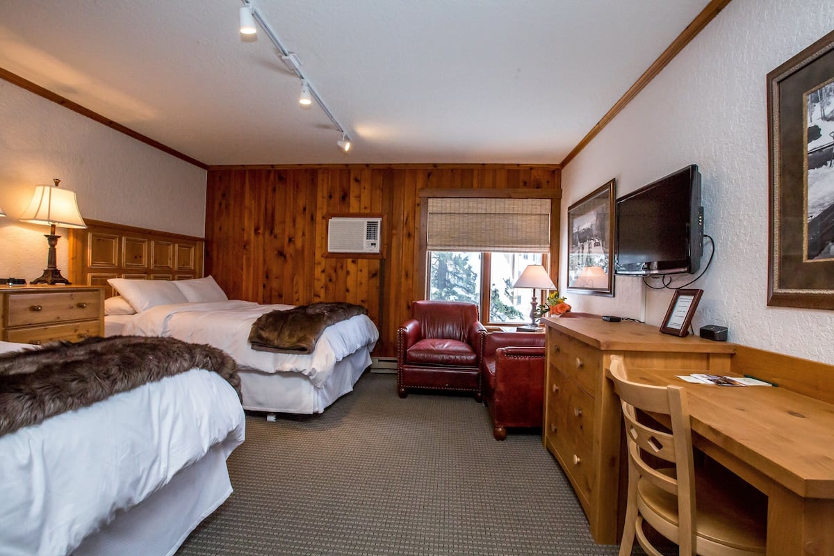 A hotel room with two beds, a desk, a chair, a television, and a window with a view of trees.