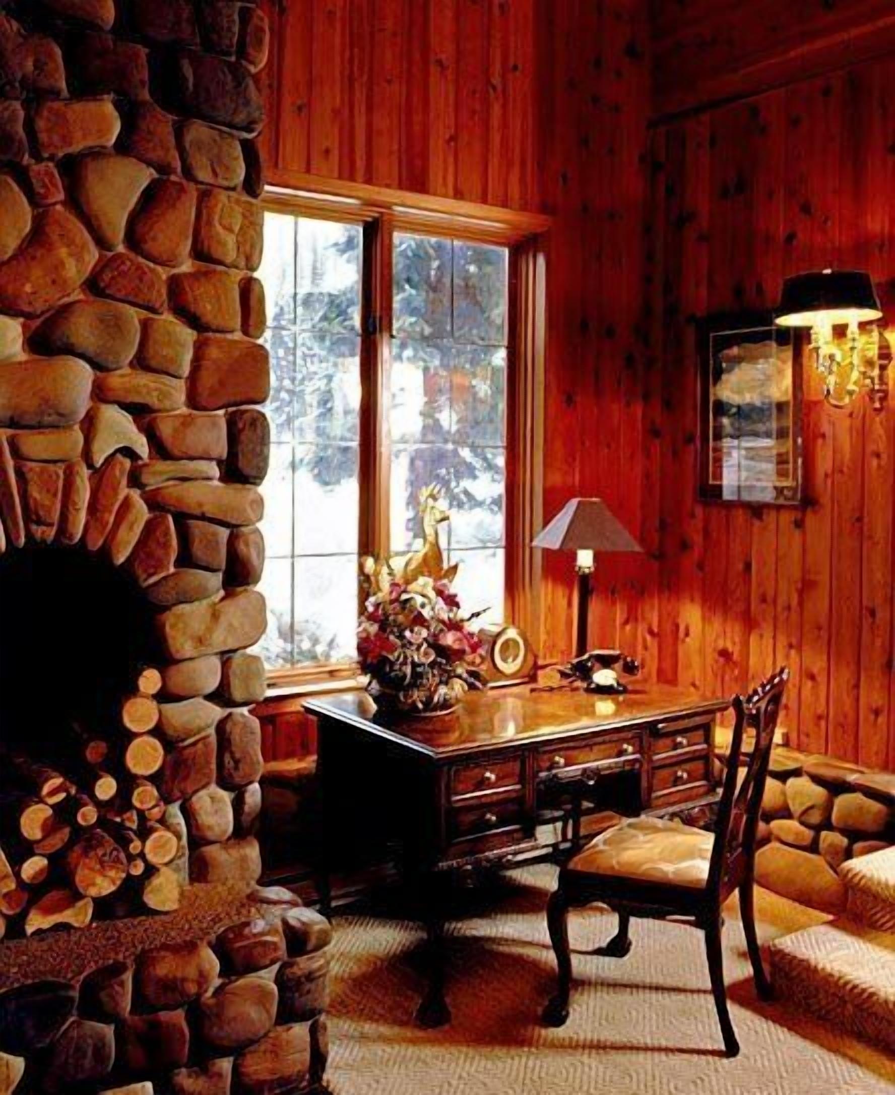 Un interior de cabaña acogedor con una chimenea de piedra, paredes de madera, un escritorio con lámpara y una ventana con vistas a árboles.