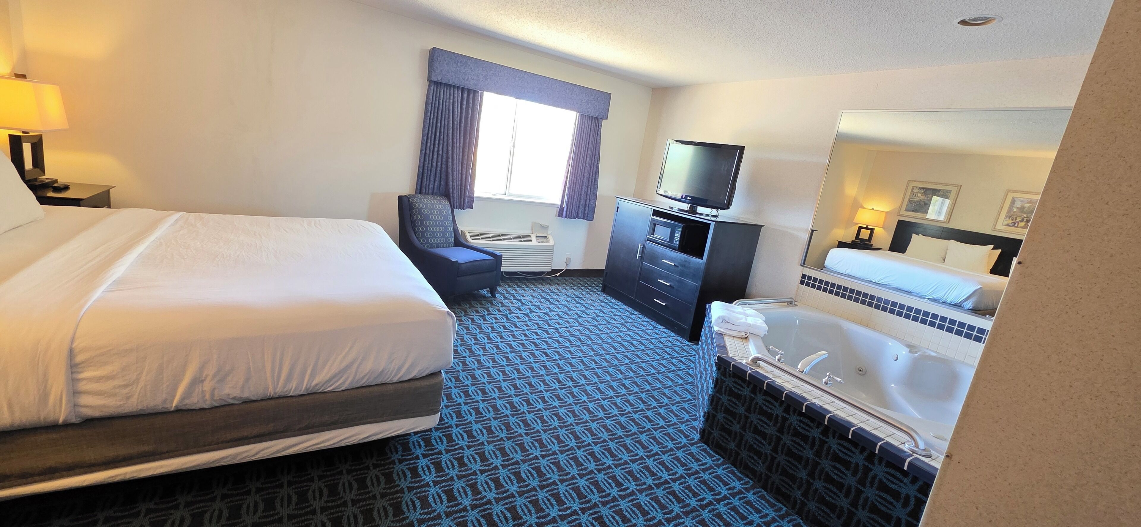 A hotel room with a large bed, a desk with a computer, a chair, a television, a microwave, a minibar, a bathroom with a bathtub, and a window with curtains.