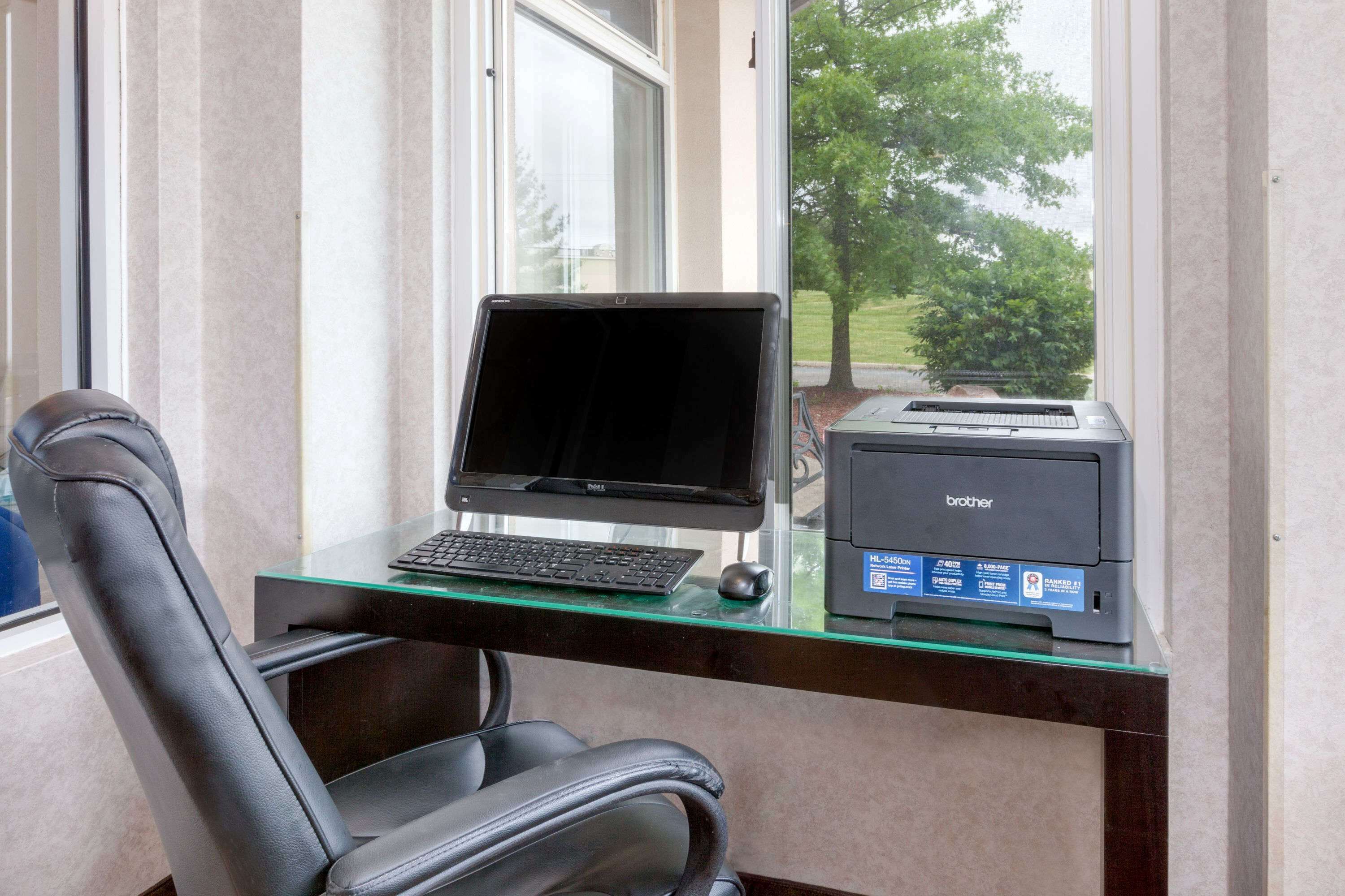Espaço de escritório em casa com computador, impressora e cadeira perto de uma janela.