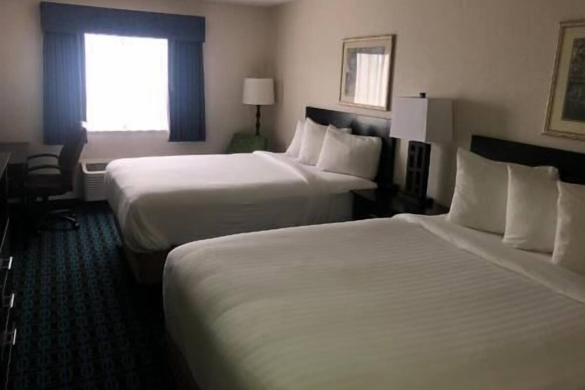 A hotel room with two beds, a desk, a chair, a lamp, and a window with curtains.