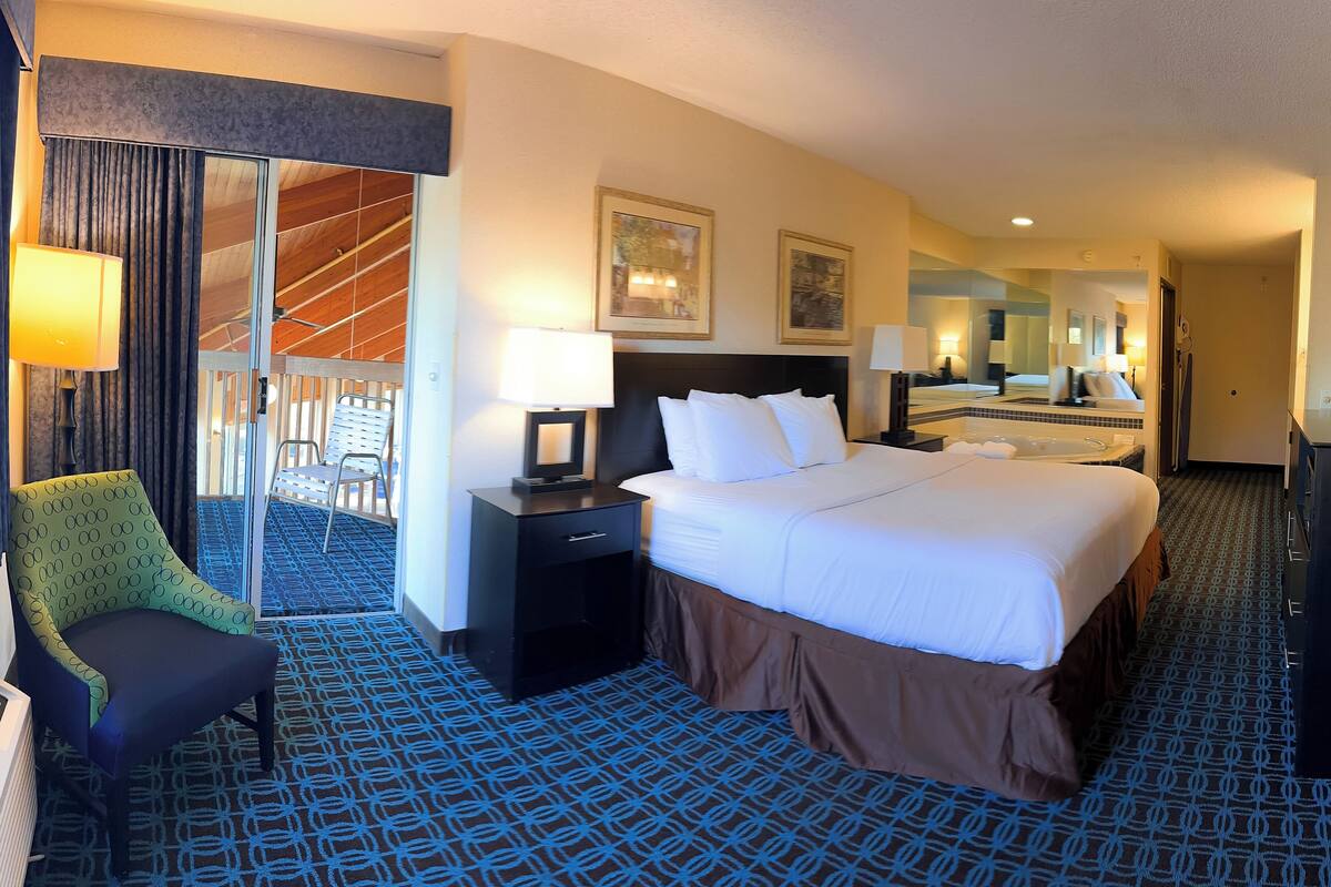 A hotel room with a large bed, two bedside tables, a desk, a chair, a bathroom with a bathtub, and a large mirror.