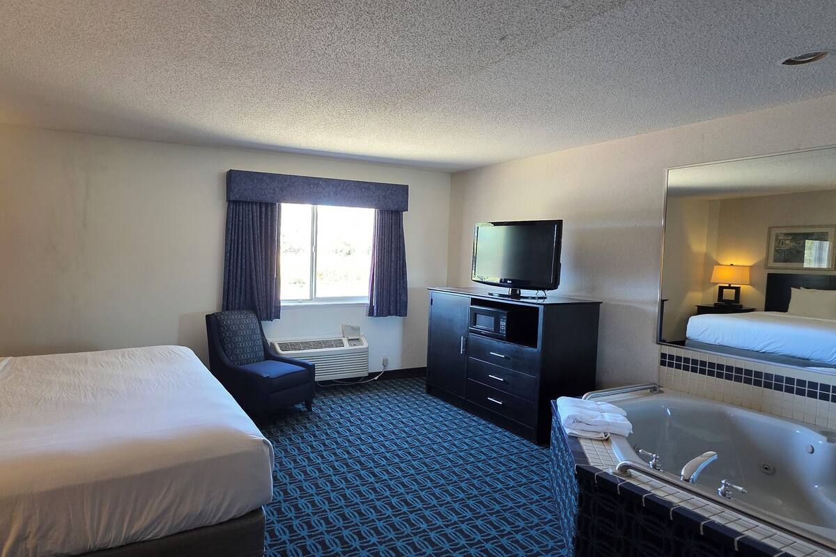 A hotel room with a bed, a desk with a lamp, a television, a desk chair, a large mirror, and a bathtub.